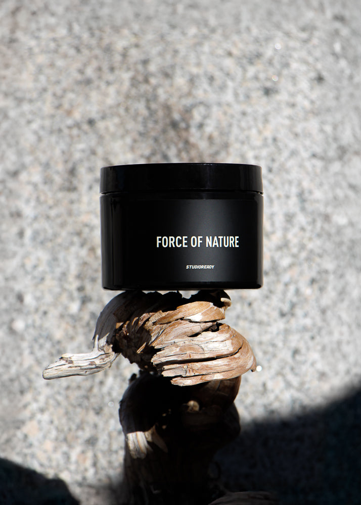 A container of Force of Nature body scrub placed on a textured surface with a shadow falling across the scene.
