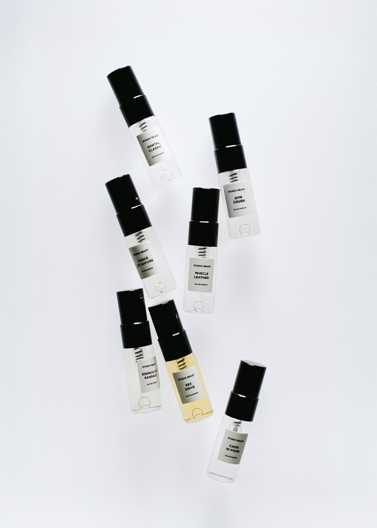 Seven pocket sized fragrance bottles with black caps arranged diagonally on a pale background