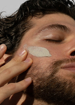 Close up of a man applying a speckled beige facial masque to his cheek with fingertips.